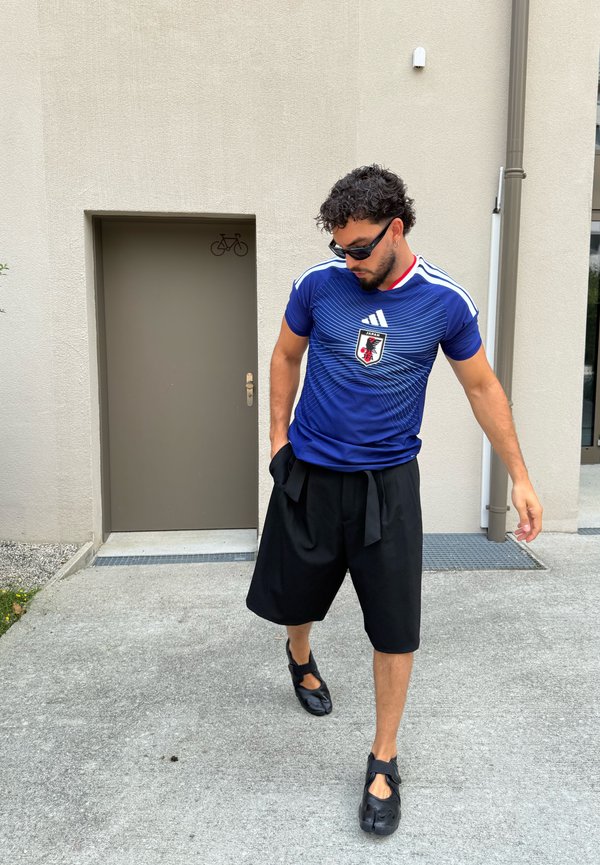 Mann trägt ein blaues Japan-Fußballtrikot, schwarze weite Shorts und schwarze Schuhe, steht draußen neben einem beigen Gebäude mit einer geschlossenen Tür.