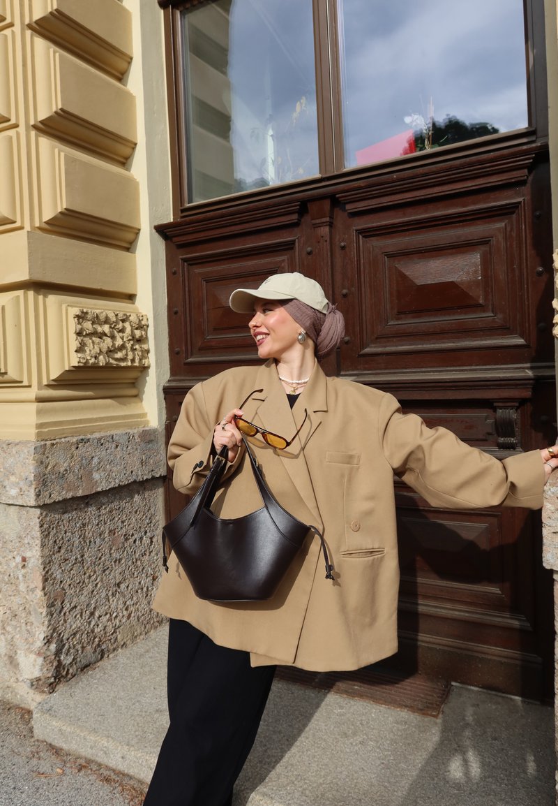 Femme portant un blazer beige oversize et une casquette, tenant un sac à main noir et des lunettes de soleil, debout près d'une porte en bois avec un mur en pierre et orné à proximité.