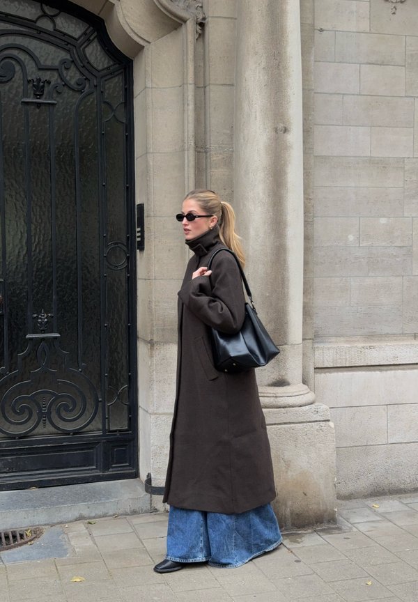 Pruun villane mantel kõrge kaelusega, sinised laiad teksapüksid ja must käekott. Lisandatud väiksed prillid. Linnaõhk taustaks.