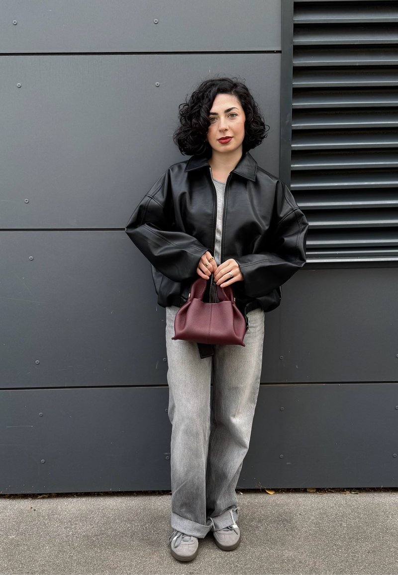 Kvinna med lockigt svart hår iförd en svart läderjacka, grå jeans, grå sneakers, som håller en liten vinröd handväska mot en grå vägg.