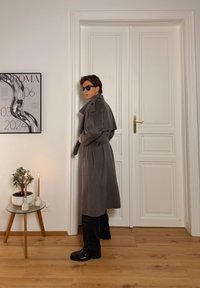 Trench-coat gris longueur genoux avec taille ceinturée et col replié, associé à un pantalon et des chaussures noirs. En arrière-plan, une plante et des bougies.