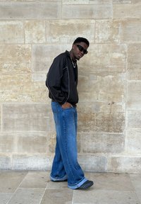 Czarna kurtka na zamek błyskawiczny z białymi akcentami, niebieskie szerokie dżinsy i czarne sneakersy. Okulary przeciwsłoneczne i łańcuszek dopełniają wygląd.