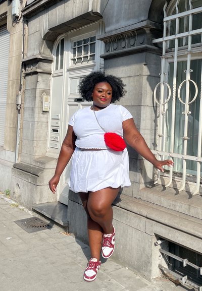 Top corto blanco y falda con un acabado texturizado. Bolso cruzado de piel sintética roja como acento. Zapatillas marrón y blancas. Caminando en un entorno urbano.