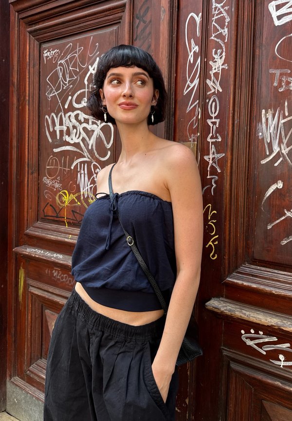 Junge Frau mit kurzen schwarzen Haaren, die ein trägerloses dunkelblaues Oberteil und schwarze Hose trägt, steht vor einer mit Graffiti bedeckten Holztür.