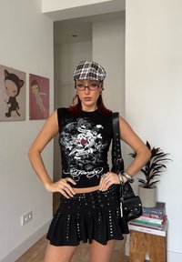Femme aux cheveux roux portant une casquette à carreaux, un haut noir sans manches avec un motif graphique, une jupe cloutée, des lunettes et des bijoux en argent, debout dans une pièce moderne avec de l'art et des plantes.