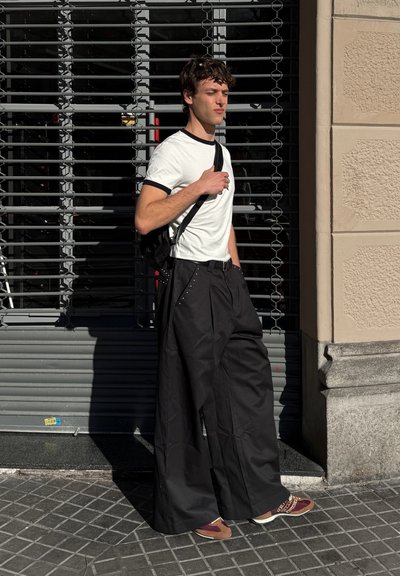 Pantalones de pierna ancha negros con acentos de tachuelas, combinados con una camiseta blanca y una mochila negra. La persona está de pie frente a un fondo de persiana metálica.