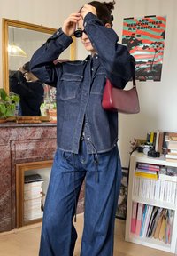 Personne ajustant des lunettes de soleil noires, portant une veste en jean foncé et un jean large, avec un sac rouge à l'épaule, debout dans une pièce avec des livres et un miroir.