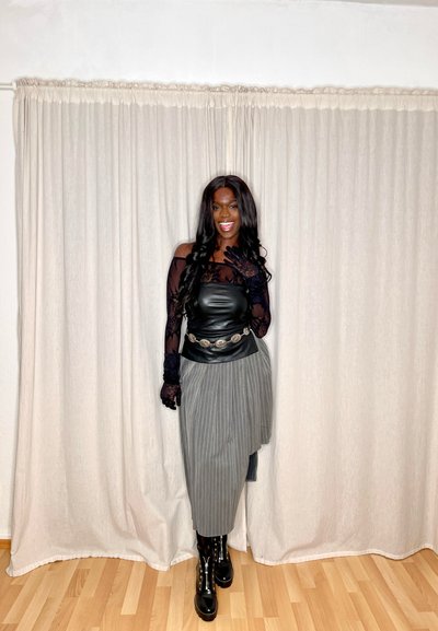 Top de cuero negro con mangas de encaje transparente, falda gris a rayas, cinturón plateado y botas de charol negras, contra un fondo de cortina.