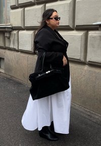 Blazer noir surdimensionné porté sur une robe blanche, accessoirisé d'un grand sac à main noir et de bottines noires. Texture lisse, design élégant.