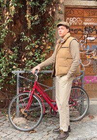 Punaisen rungon ja mustan korin sisältävä polkupyörä, joka on asetettu henkilön viereen. Henkilöllä on päällään vaaleanruskea liivi, beige housut ja tummat kengät, taustana vihreä maisema.