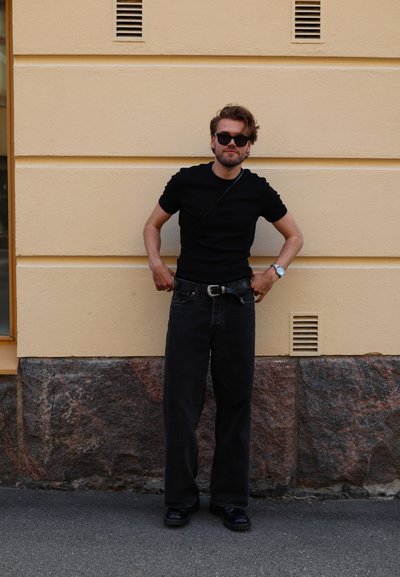 Hombre con gafas de sol negras, camiseta negra y jeans negros, ajustándose el cinturón mientras está de pie contra una pared beige y de piedra.