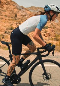 Ciclista in maglia azzurro chiaro e bianca, pantaloni neri e scarpe da ciclismo, che pedala su un sentiero roccioso su una bicicletta nera, mostrando tatuaggi sulle gambe.