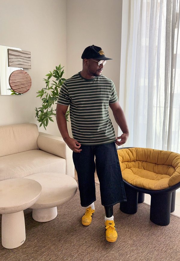 Mężczyzna o ciemnej karnacji, ubrany w pasiaste ubranie, czapkę, okulary przeciwsłoneczne, szerokie czarne spodnie, żółte buty i protezę nogi, stojący w nowoczesnym salonie.