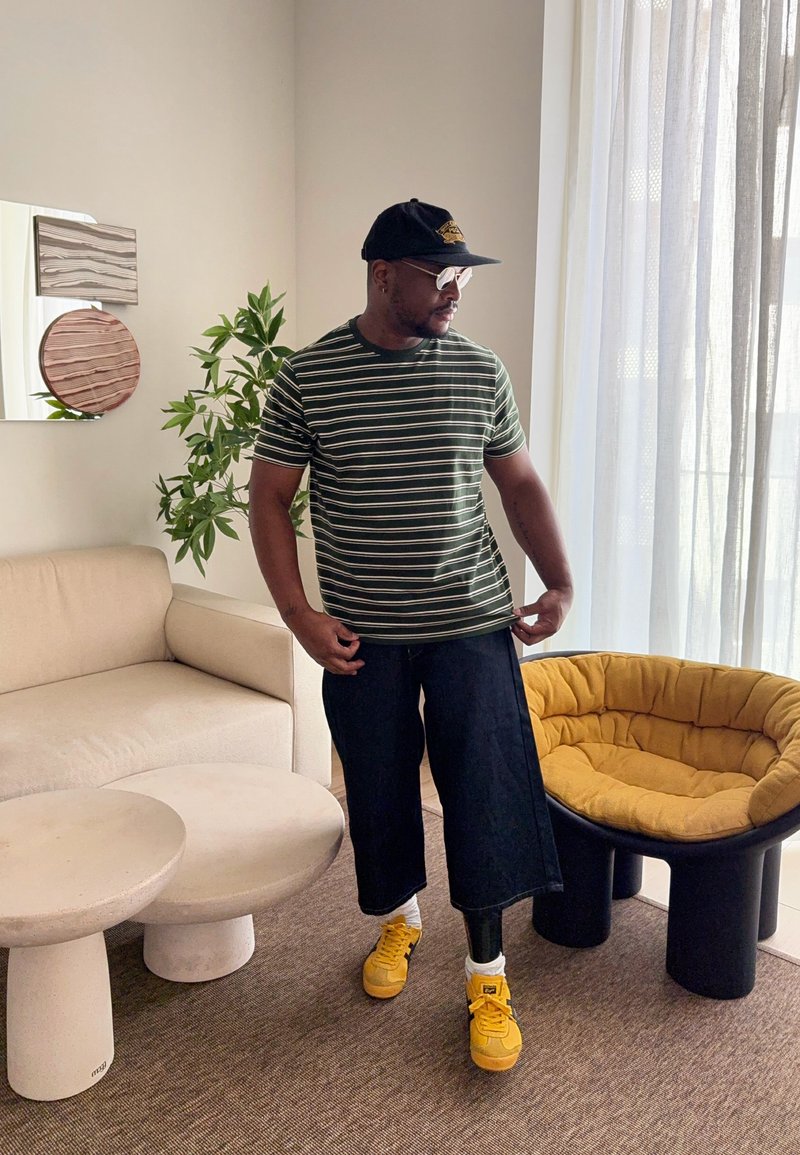 Homme à la peau foncée portant une chemise rayée, une casquette, des lunettes de soleil, un pantalon large noir, des chaussures jaunes et une jambe prothétique, debout dans un salon moderne.