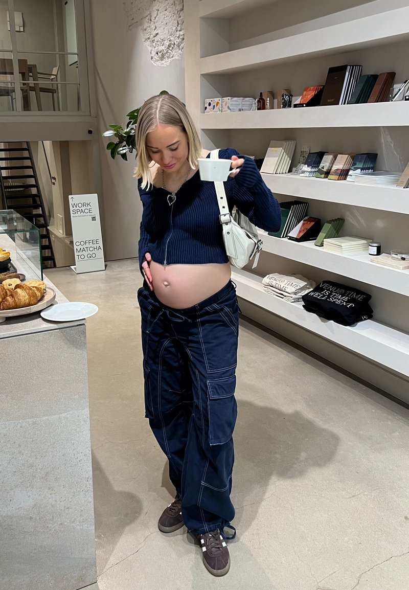 Σκούρο μπλε φούτερ με φερμουάρ, cargo παντελόνι με πολλές τσέπες και σκούρα αθλητικά παπούτσια. Κρατάει ένα φλιτζάνι ενώ αγγίζει μια στρογγυλή κοιλιά σε ένα μοντέρνο καφέ.