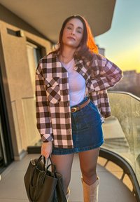 Černobílá kostkovaná flanelová košile, pod ní bílý tričko, modrá denimová sukně, hnědý opasek, drží černou kabelku, na nohou světle hnědé kozačky.