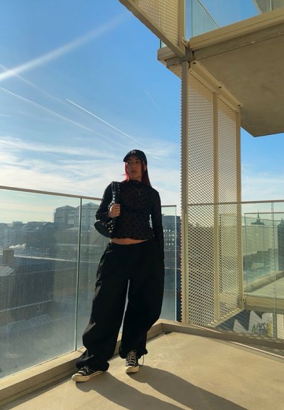 Top de manga larga negro con lunares, pantalones anchos negros, zapatillas negras y una gorra negra. De pie en un balcón con vista a la ciudad.