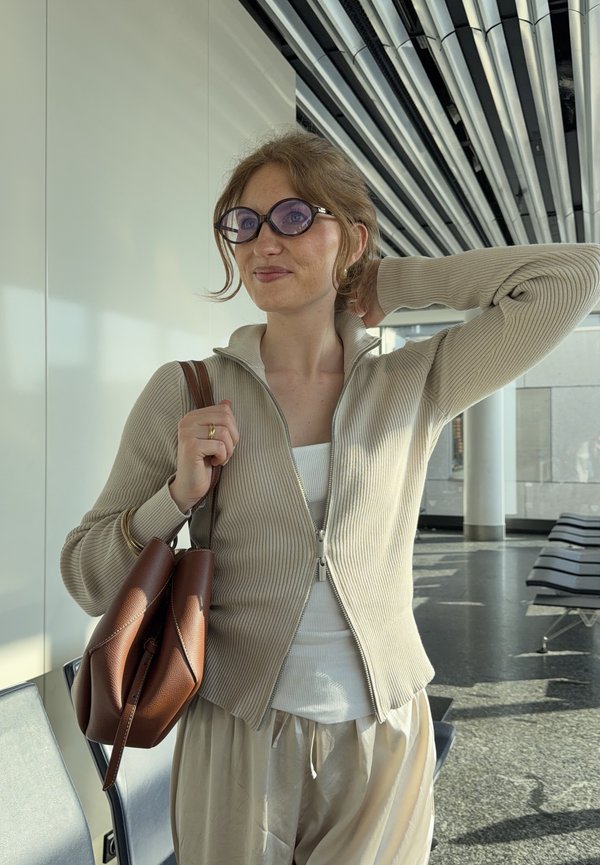 Joven mujer con gafas y ropa beige sosteniendo un bolso de cuero marrón en una zona de espera moderna con sillas de metal y techo a rayas.