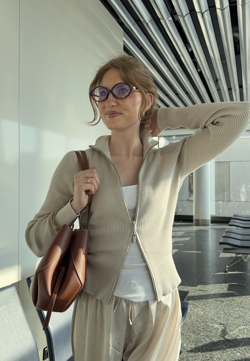 Jeune femme portant des lunettes et des vêtements beige tenant un sac en cuir marron dans une salle d'attente moderne avec des chaises en métal et un plafond rayé.