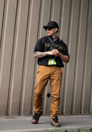 Camisa negra con un diseño gráfico en amarillo y verde, pantalones beige y zapatillas negras con detalles marrones. El fondo presenta líneas verticales de hormigón.