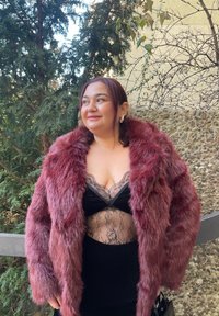 Manteau en fausse fourrure bordeaux avec col châle, porté sur un body en dentelle noire. Robe noire en dessous, sur fond naturel.