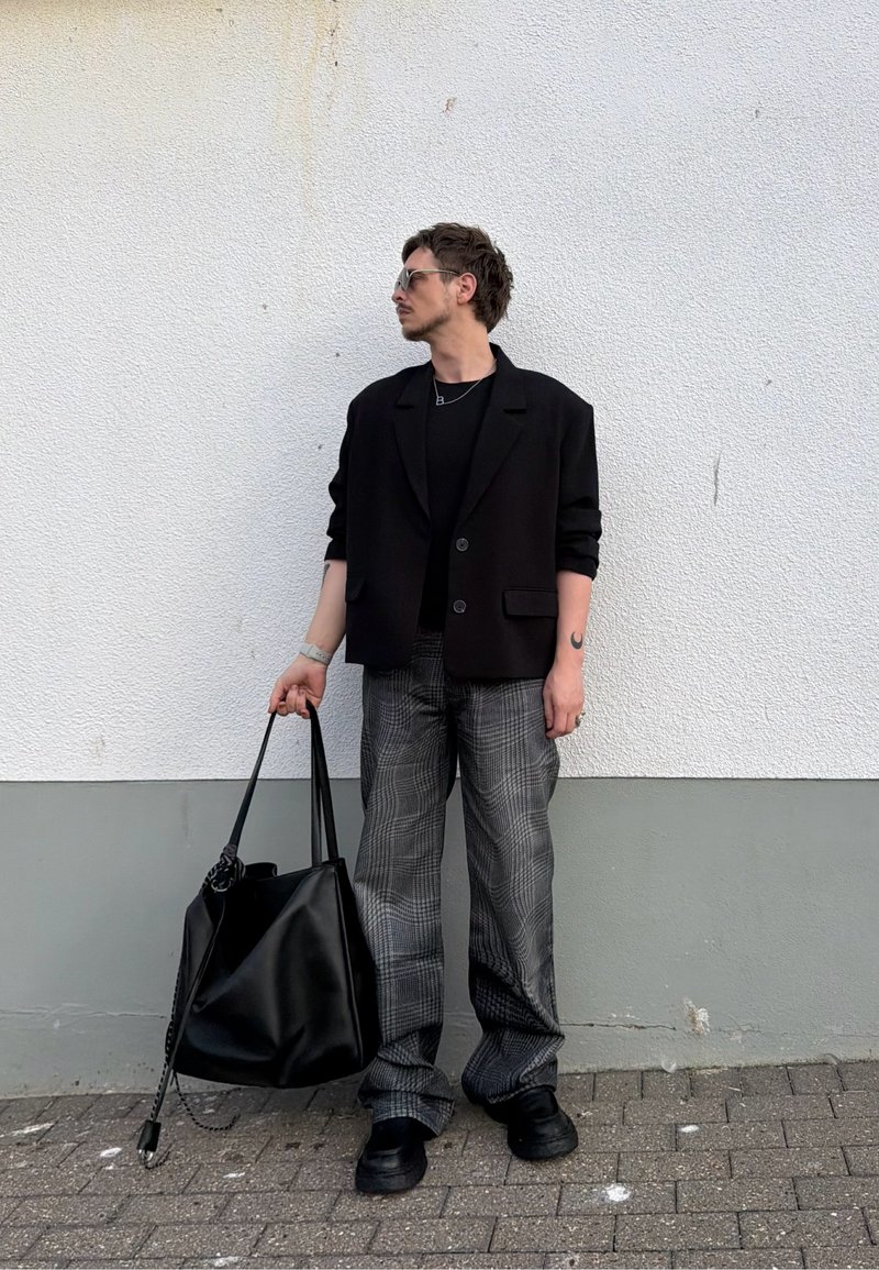 Mann iført svart blazer og rutete bukser står mot en hvit vegg, holder en stor svart veske i venstre hånd og ser mot høyre.