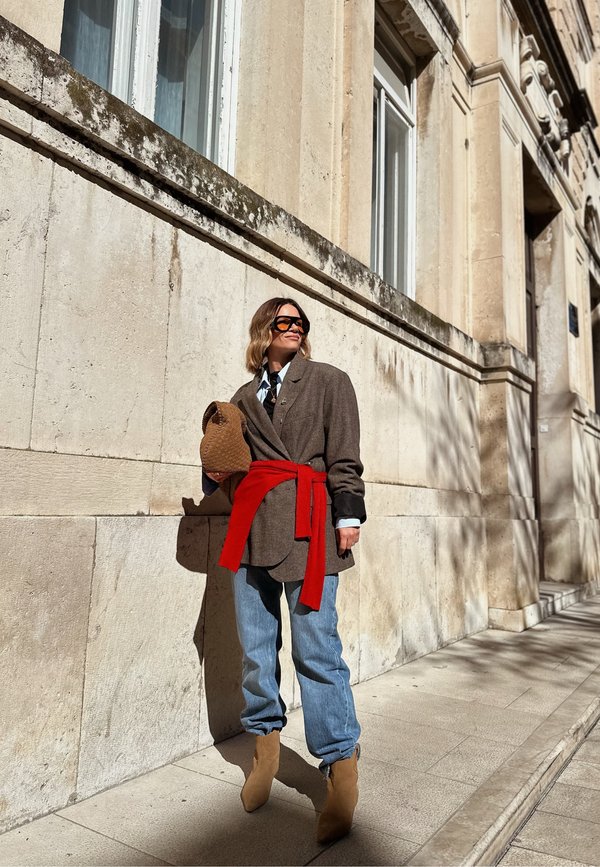 Vrouw draagt een oversized bruine blazer, rode trui gestrikt rond de taille, lichtblauwe spijkerbroek, beige laarzen, houdt een bruine geweven tas vast, staat bij een stenen gebouwmuur.