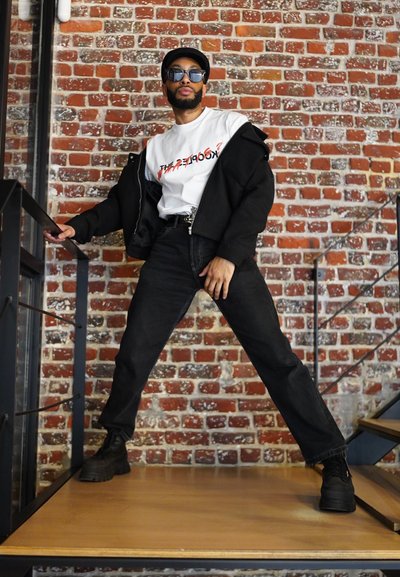 Chaqueta negra de gran tamaño superpuesta sobre una camiseta gráfica blanca, combinada con jeans oscuros y botas negras gruesas. De pie sobre una plataforma de madera.