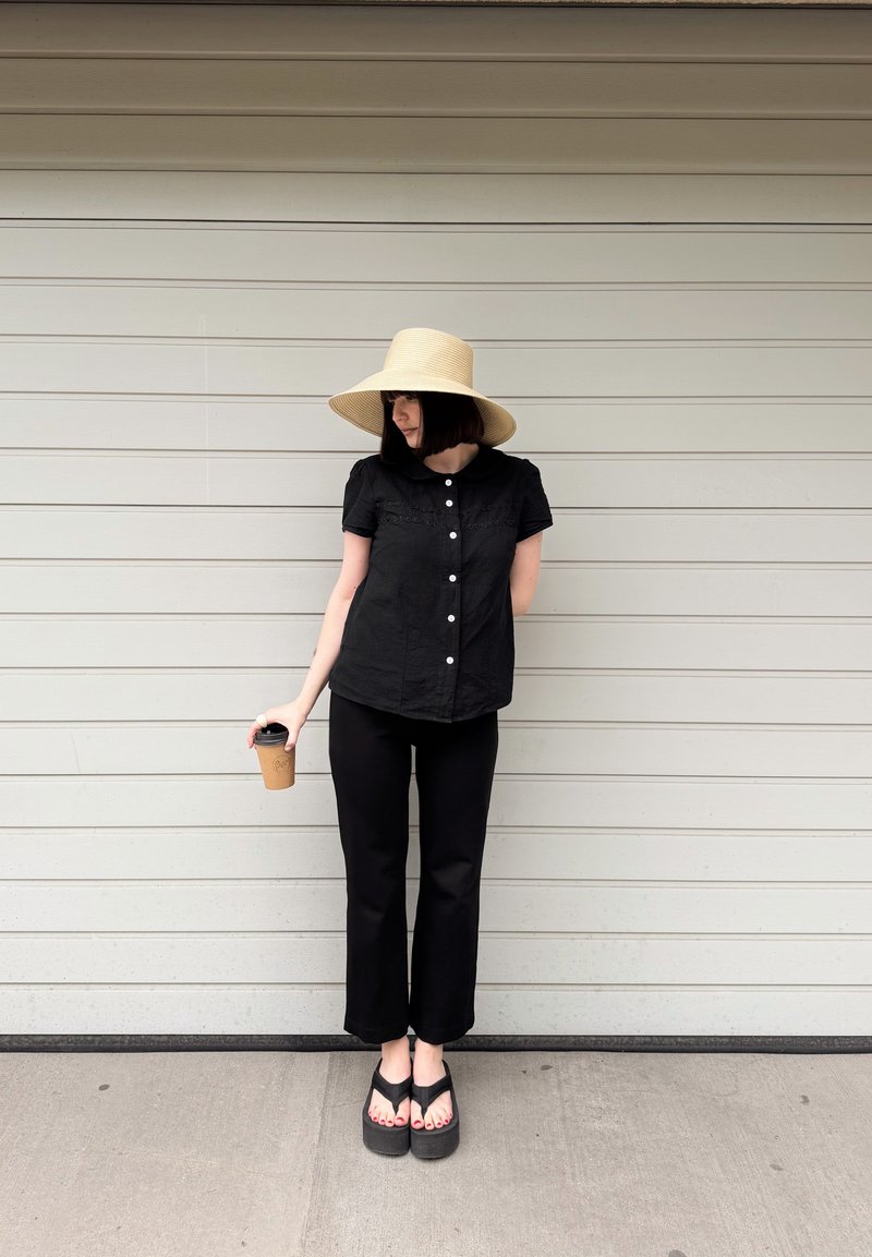 Femme en tenue noire et chapeau à larges bords, debout contre un mur gris clair, tenant une tasse à café à emporter dans sa main droite.
