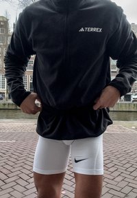 Pull-over en polaire noire avec fermeture éclair de Terrex ; shorts de compression blancs avec logo Nike noir. Les deux matériaux semblent doux et ajustés.