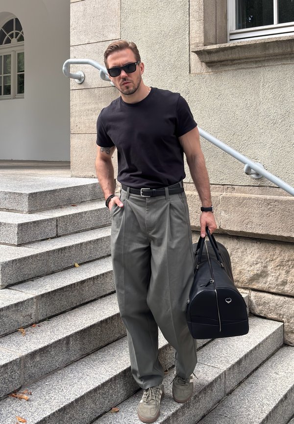 Uomo con occhiali da sole, t-shirt nero e pantaloni grigi, che porta una borsa da palestra nera mentre scende una scalinata di pietra fuori da un edificio.