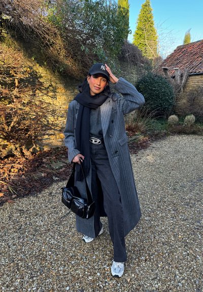 Abrigo gris a rayas sobre un suéter negro y jeans. Lleva un bolso negro; usa zapatillas blancas y una gorra negra. Entorno al aire libre con vegetación.