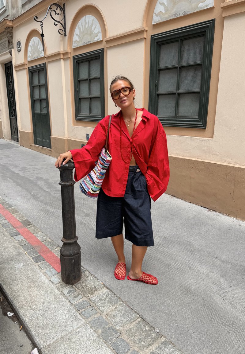 Femme en blouse rouge, short noir jusqu'aux genoux et chaussures rouges, appuyée contre un poteau en métal sur un trottoir en ville avec un bâtiment beige et des fenêtres derrière elle.