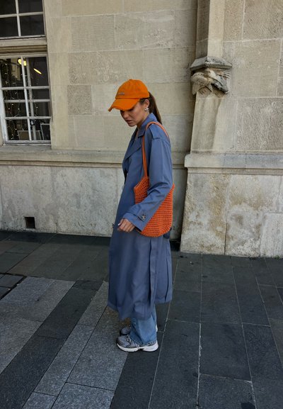 Mujer con gorra y bolso naranja, gabardina azul, vaqueros y zapatillas caminando por la acera cerca de un edificio de piedra con una ventana y tallados decorativos.