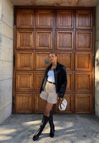 Donna che indossa una giacca di pelle nera, una maglietta bianca, pantaloni corti beige e stivali neri al ginocchio, tiene in mano una piccola borsa bianca, in piedi davanti a una porta in legno a pannelli.