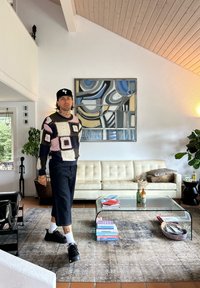 Mies, joka käyttää kuvioitua villapaitaa ja myssyä, kävelee modernissa olohuoneessa, jossa on valkoinen sohva, abstrakti maalaus ja lasinen kahvipöytä, jossa on kirjoja.