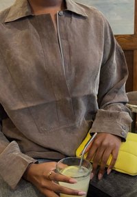 Eine Person in einem grauen Knopfhemd hält ein Glas mit Zitronenwasser und einem Metallstrohhalm, während sie die Hand auf einer leuchtend gelben gesteppte Tasche resting.