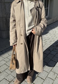 Osoba v béžovém trenčkotě, krémovém pleteném svetru, tmavých kalhotách a béžových botách, držící hnědou kabelku, stojí na kamenném chodníku.