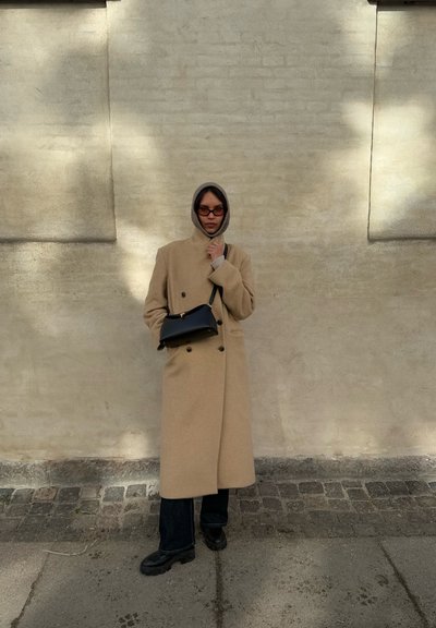 Abrigo de lana beige con diseño de doble botonadura, llevado sobre una sudadera gris con capucha. Bolso negro. Jeans oscuros y botas negras gruesas completan el atuendo.