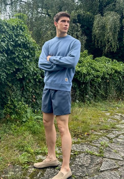 Sudadera azul con cuello redondo y puños de canalé, combinada con pantalones cortos azul marino. Zapatillas beige sin cordones, de pie sobre un camino de piedras rodeado de vegetación.