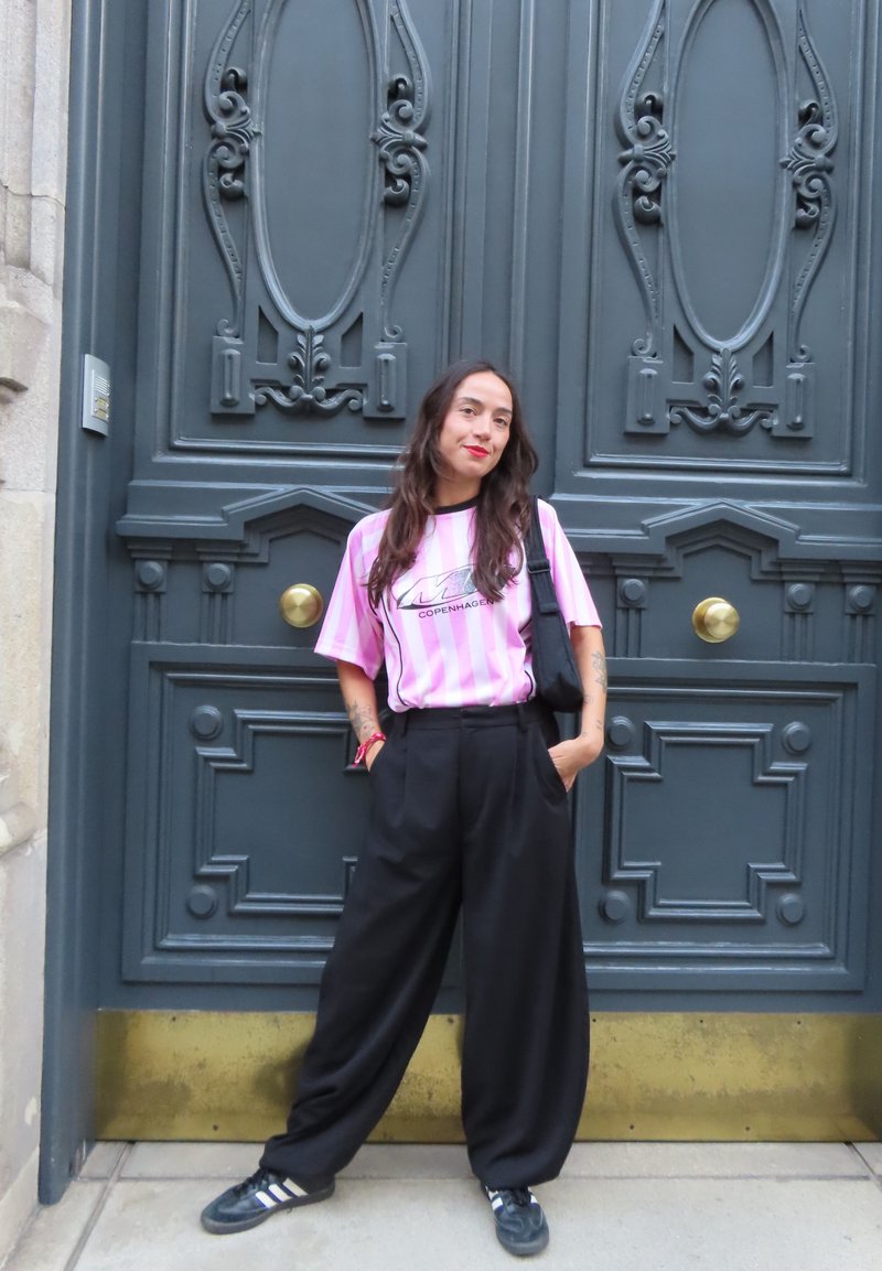 T-shirt às riscas rosa e branca com estampado gráfico, calças largas pretas e ténis pretos, em pé contra uma porta decorativa escura.