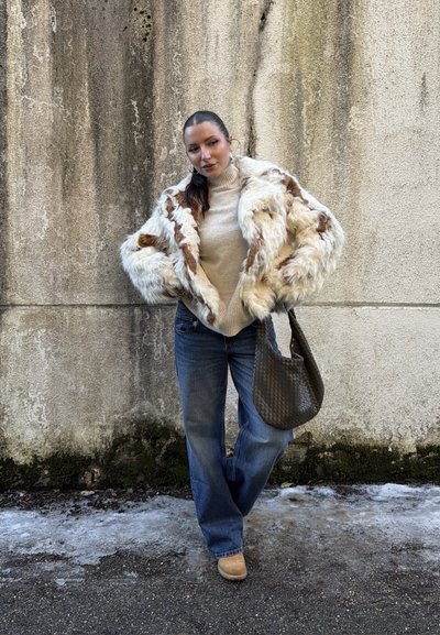 Mujer con chaqueta de piel blanca y marrón, cuello alto color crema, vaqueros azules, llevando un bolso grande oscuro con textura, de pie sobre suelo helado junto a una pared de hormigón.