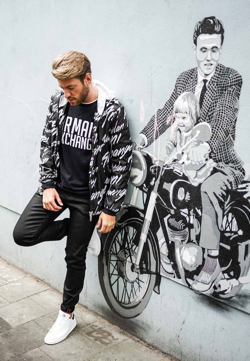 Black and white zip-up jacket with abstract text pattern, worn over a dark shirt. Black pants with white sneakers, posed beside mural of a man and child on a motorcycle.