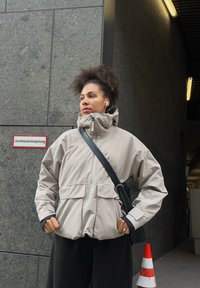 Noor curly juustega noor naine, kes kannab beeži jakk ja musta õlakotiga, seisab halli seina ja traffic cone'i lähedal linnapildis.