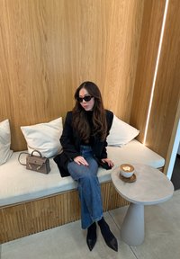 Ένα καθιστό άτομο φορά μαύρα γυαλιά ηλίου, μαύρη μπλούζα και μια μακριά ζακέτα με μπλε τζιν. Δίπλα τους υπάρχει μια γκρίζα τσάντα, ενώ σε ένα στρογγυλό τραπέζι βρίσκεται καφές.