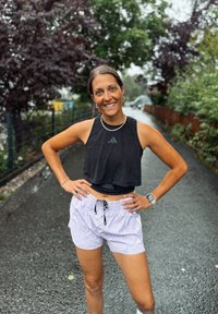 En smilende kvinne i svart ermeløs topp og lyse lilla shorts står med hendene på hoftene på en fuktig utendørssti omgitt av trær og rekkverk.