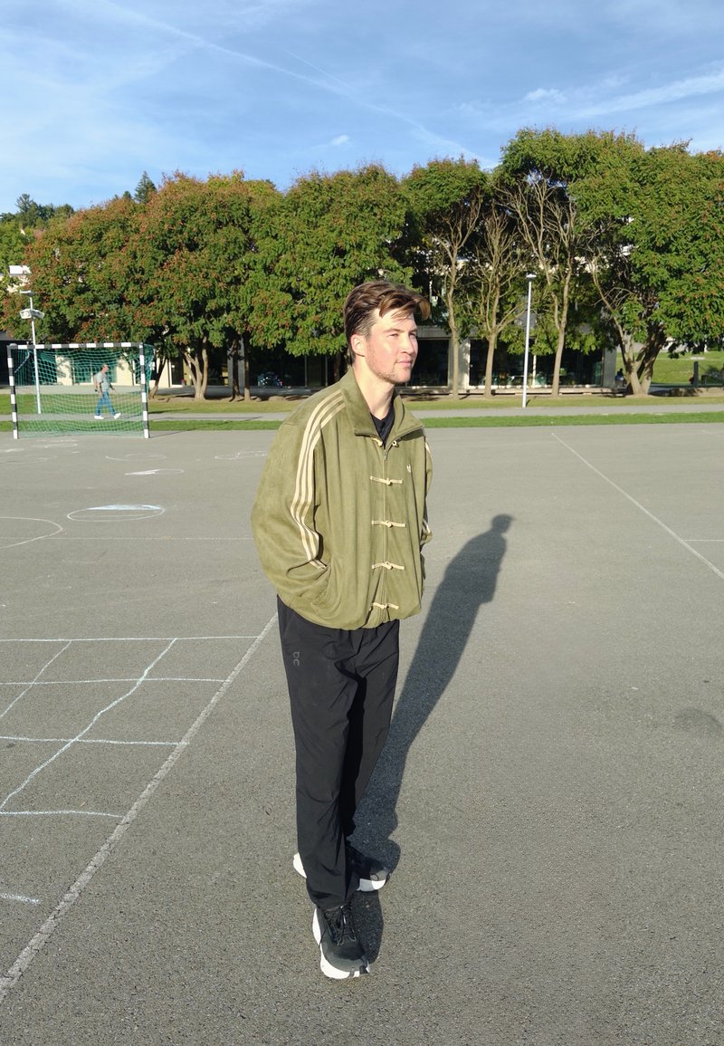 Hombre con chaqueta verde y pantalones negros de pie en una cancha deportiva pavimentada con árboles y una portería de fútbol al fondo.