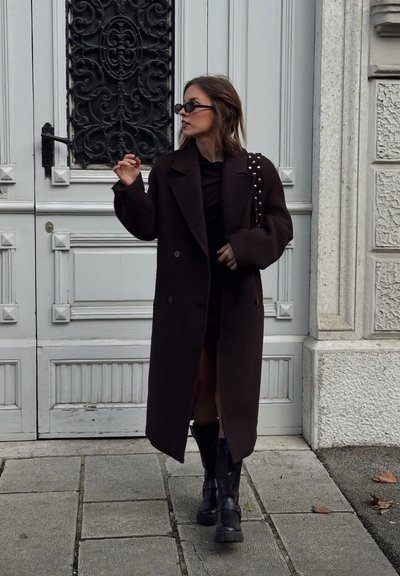 Mujer con un abrigo largo de color oscuro, botas negras y gafas de sol, de pie sobre un pavimento de piedra frente a una gran puerta blanca ornamentada.