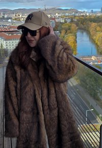 Barna műszőrme kabát plüss tapintású, bő szabású és látható varrásokkal. Párosítva bézs baseball sapkával és sötét napszemüveggel.