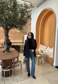 Μαύρο σακάκι πάνω από ένα μαύρο τζέρσεϊ, ψηλόμεσες μπλε τζιν, μυτερές μαύρες γόβες, γκρίζα τσάντα; τοποθετημένο κοντά σε ένα ξύλινο τραπέζι και ένα δέντρο.